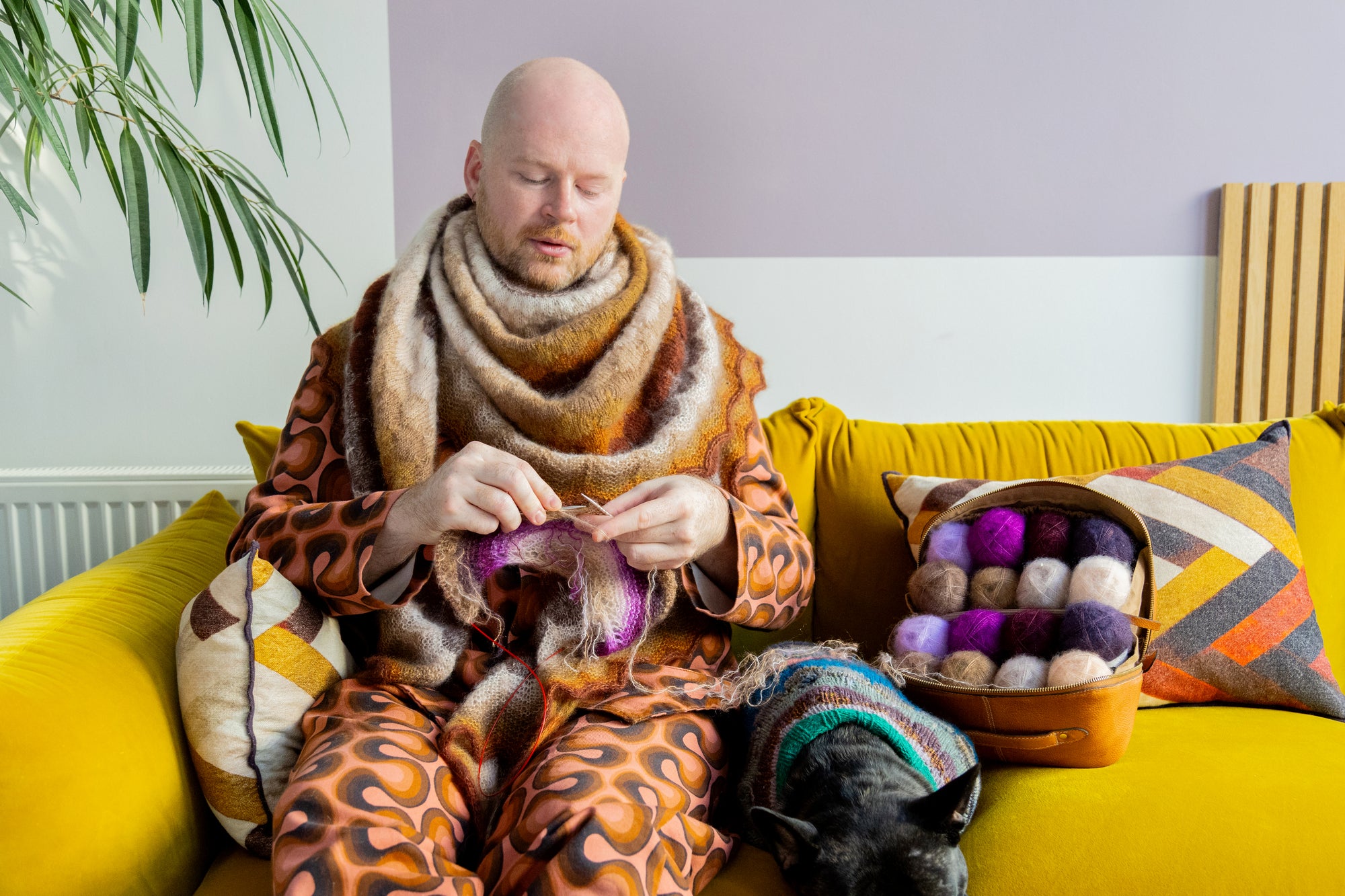 This image shows designer Stephen West wearing a beautifully knitted shawl around his neck. He is sitting on a bright yellow sofa and he is knitting a project. Next to him, lay a small dog wearing a knitted sweater and a Re:Designed open bag filled with yarn. 