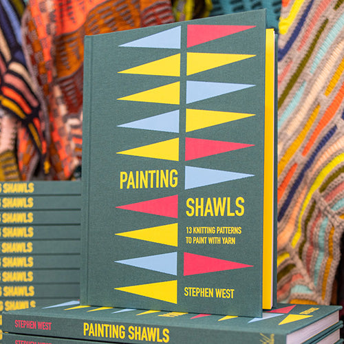 Stack of Painting Shawl books by Stephen West. Green books piled up and stools with a knitted shawls brightly displayed in the background. 