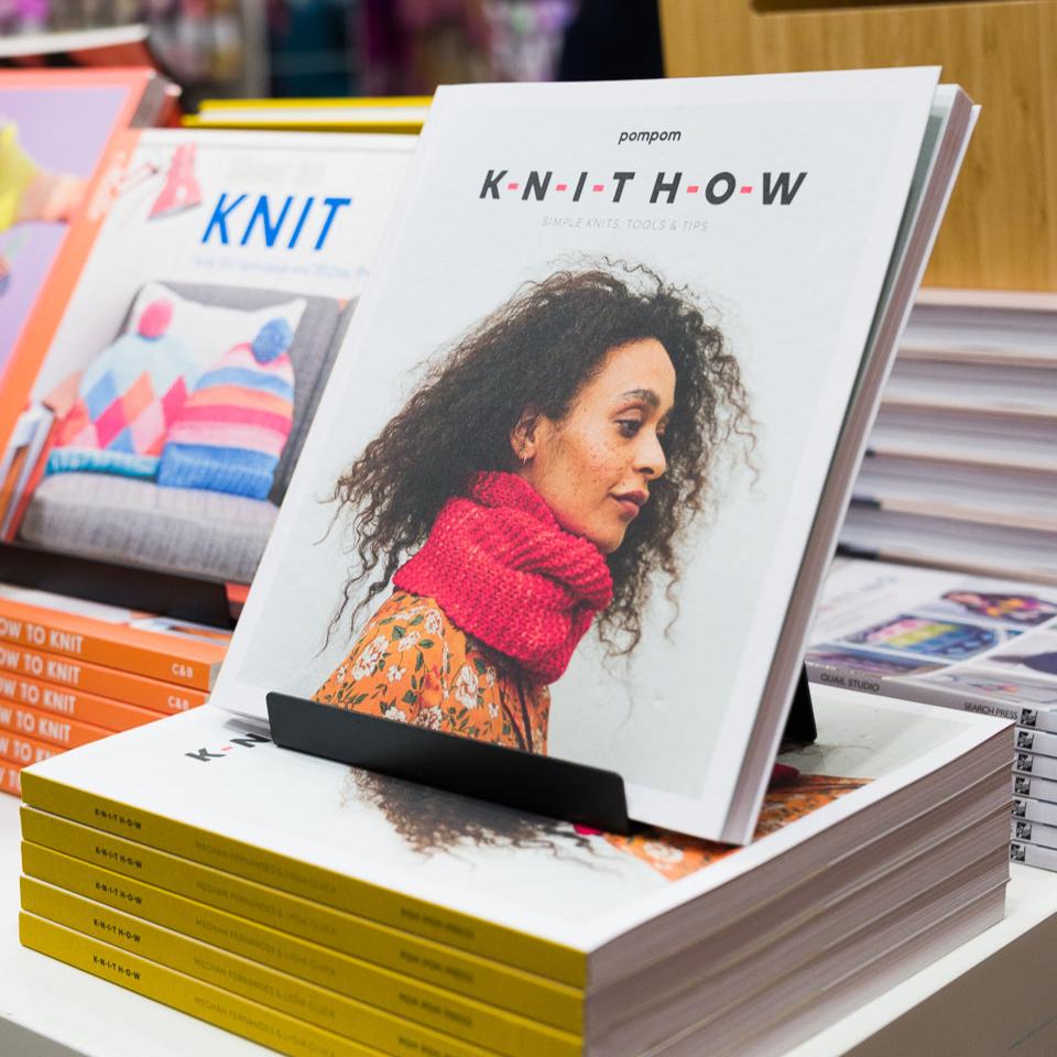 Knithow book from Pom Pom displayed in store on a pile of books. The cover image is of a person with curly hair and freckles wearing a pink knitted scarf. 