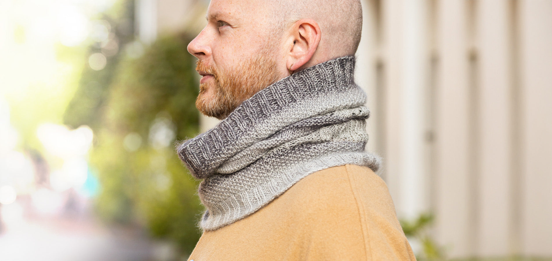 Stephen West Man standing with his profile to camera in a outside blurry setting wearing a handknitted cowl neck scarf in shades of grey