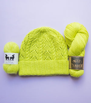 Flat lay image of a knitted beanie hat n green pickle juice colour with a ball kof West Wool mohair and skein of DK merino wool 