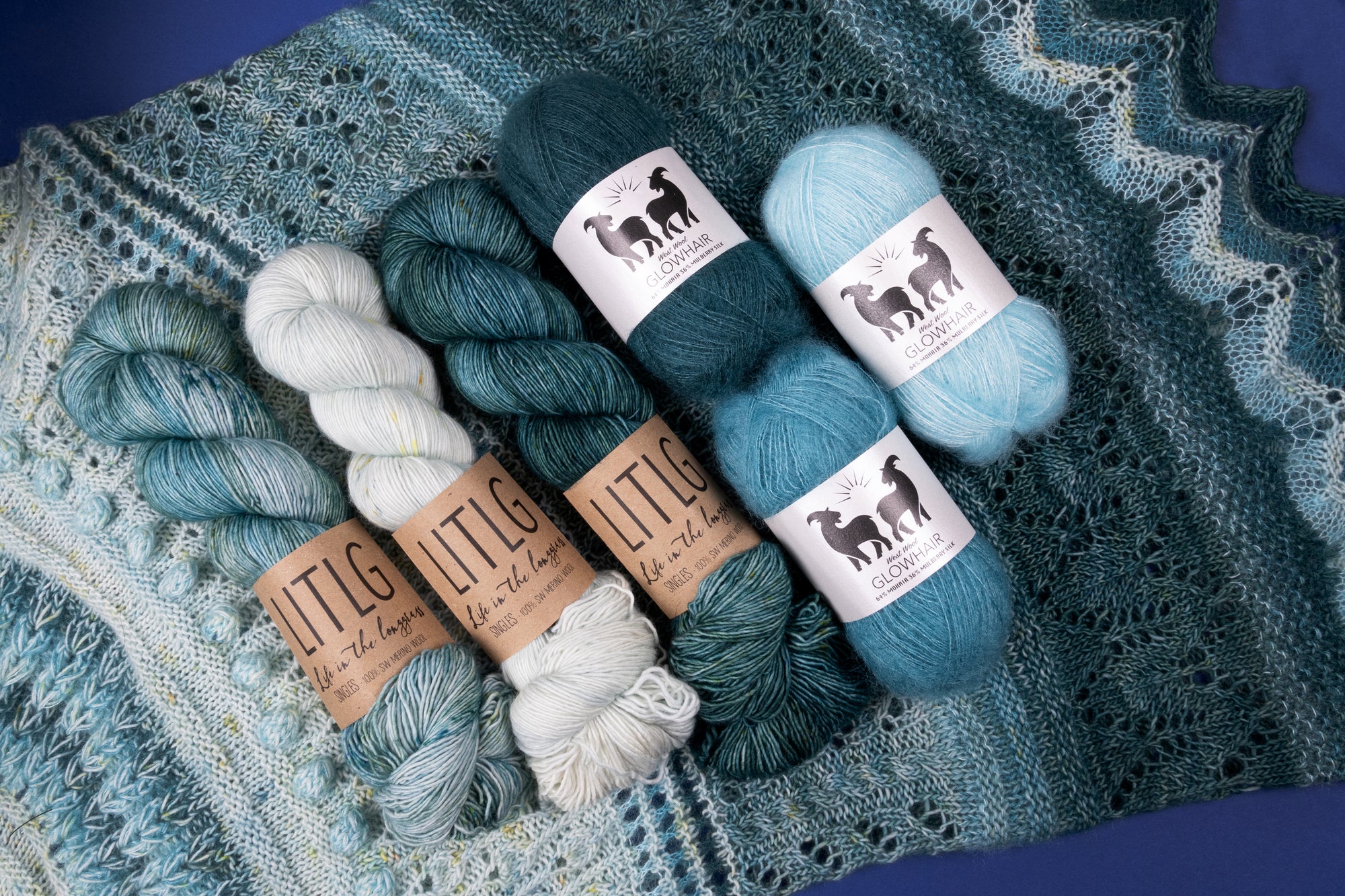 Flat lay of yarn skeins in teal greens and blues, and frosty white tones, labeled 'LITLG' and 'Glowhair,' arranged on a textured, teal-toned knitted shawl from Stephen West of West Knits. A cozy textured image.