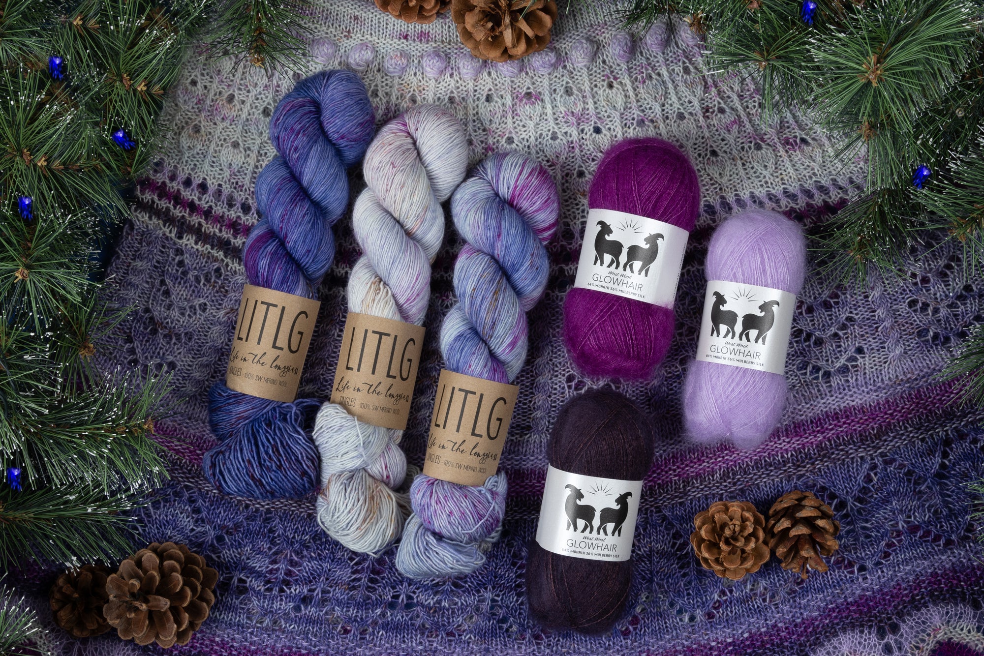 Flat lay of yarn skeins in purple, lavender, and cream tones, labeled 'LITLG' and 'Glowhair,' arranged on a textured, purple-toned knitted shawl from Stephen West of West Knits. Surrounding the yarn are sprigs of pine with small blue ornaments and several pinecones, creating a cozy, festive look.