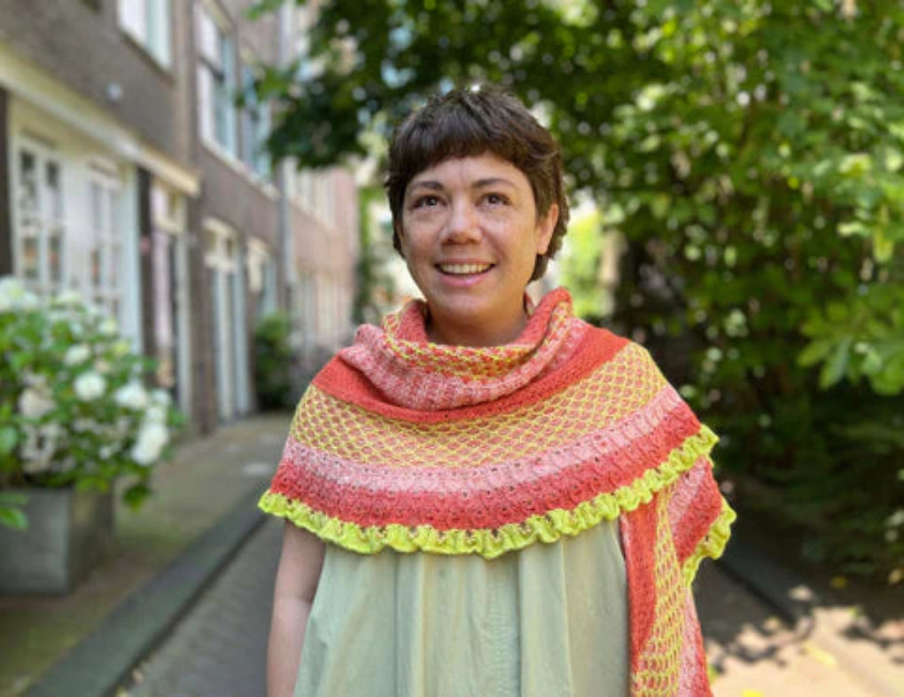 This image shows designer Malia Joseph wearing a beautifully knitted shawl in warm, poppy tones, including yellow and pink. The background shows a street with trees and houses on each side. The shawl is named "Stitchscape Shawl" and it was originally designed for the 2024 Yarn Along. 