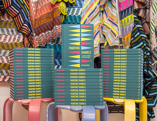 Stack of Painting Shawl books by Stephen West. Green books piled up and stools with a knitted shawls brightly displayed in the background. 