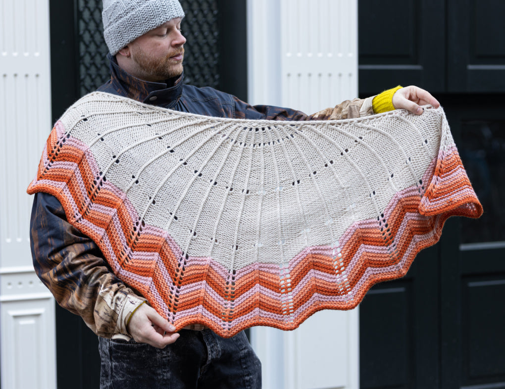 The image shows Stephen West holding the Retro Chevron Shawl. The shawl is knitted with cream, orange, and pink tones. The background shows a house with a dark door. 