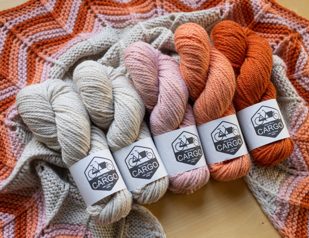 This image shows 5 skeins of chunky weight Cargo in cream, pink, and orange tones. They stand on a beautifully knitted sample of the Retro Chevron Shawl in the same colour combination. 