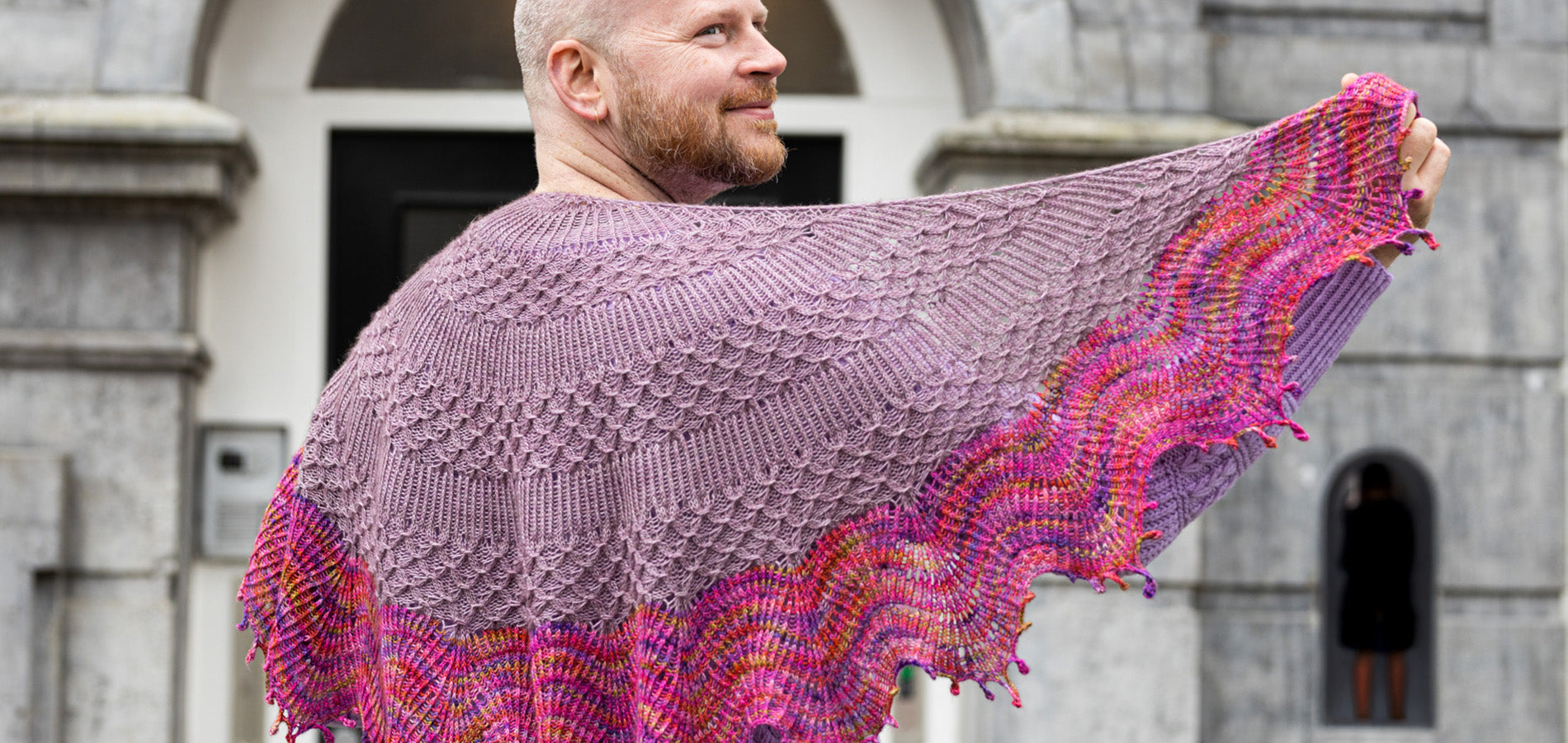 Stephen West Man standing with his back  to camera infront of a stone building wearing a handknitted shawl scarf in shades of purple and pinks and orange. 