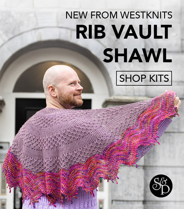 Stephen West Man standing with his back  to camera infront of a stone building wearing a handknitted shawl scarf in shades of purple and pinks and orange. 

Text reads New from west knits Rib Vault Shawl Shop Kits