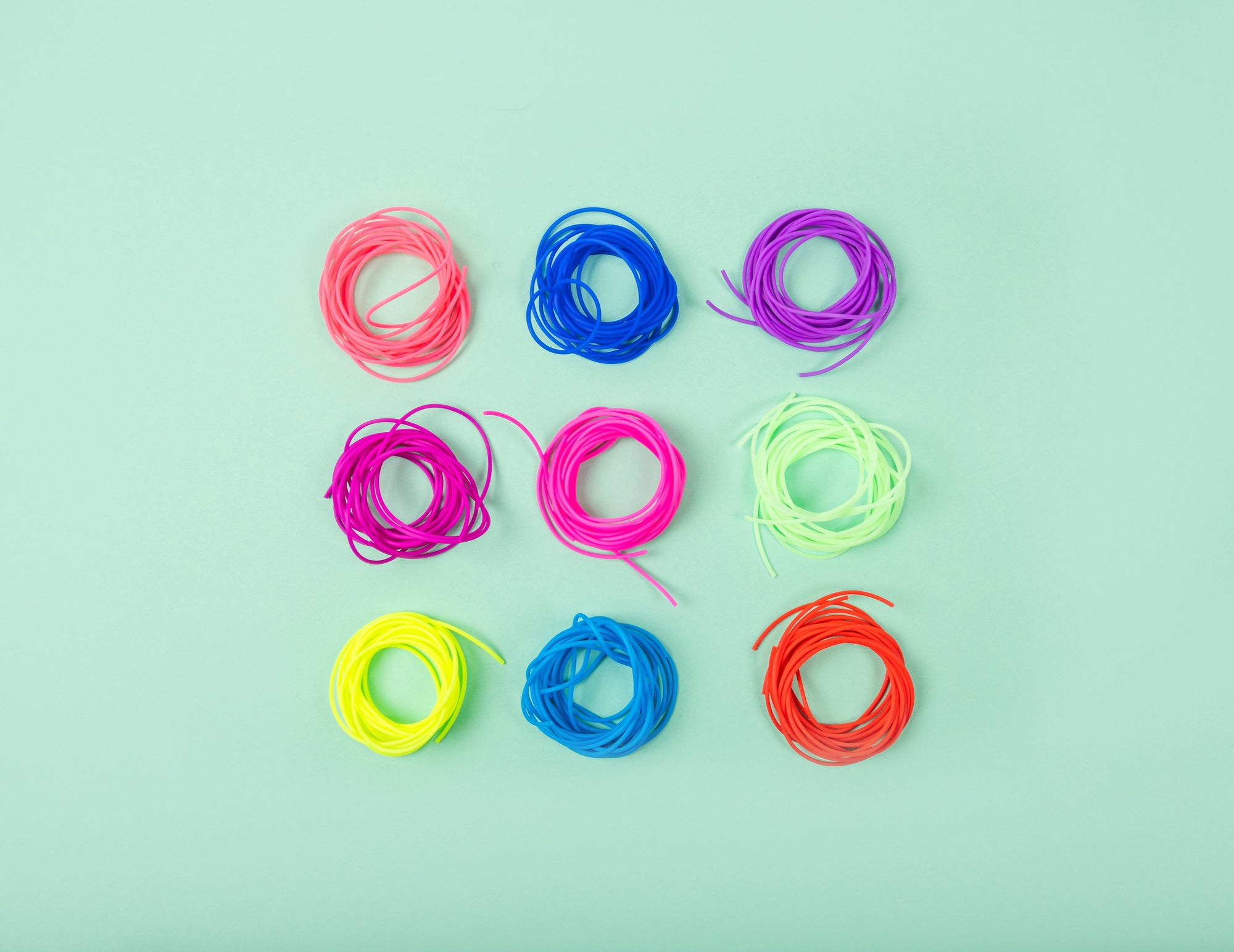 This image shows needles cords from the brand The Knitting Barber. Each cord is arranged in a circular shape, and they are displayed together in a square layout. The cords come in various bright colours, and the background is light green. 