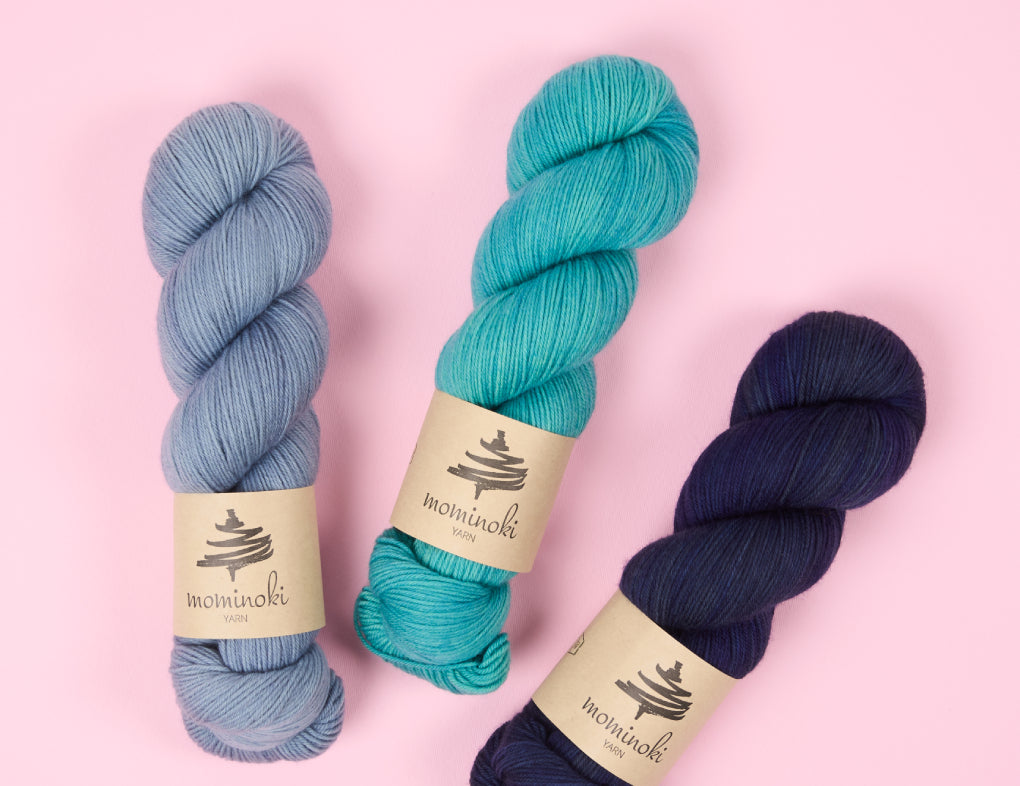 This image shows three skeins of Sock Fine 4ply from the brand Mominoki Yarn. The skeins are in three shades of blue and they stand on a pastel pink background. 