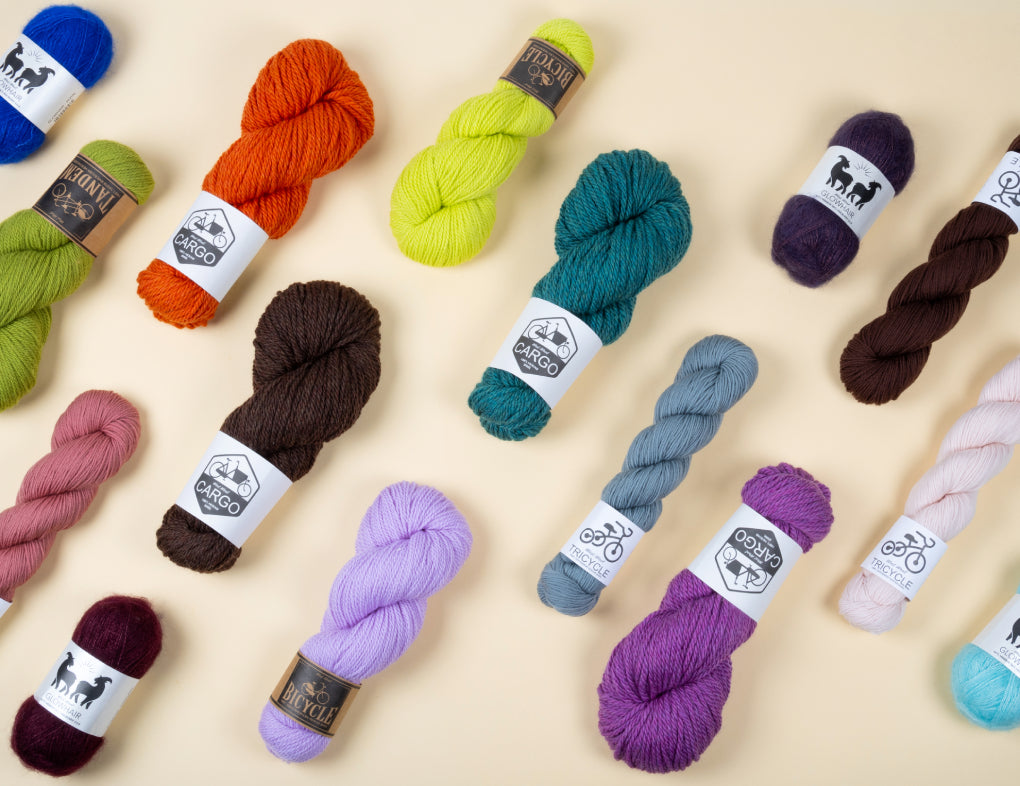 This image shows various skeins of the brand West Wool on a pastel yellow background. The skeins are in various colours and bases (Tricycle, Tandem, Bicycle, Glowhair, Cargo). 