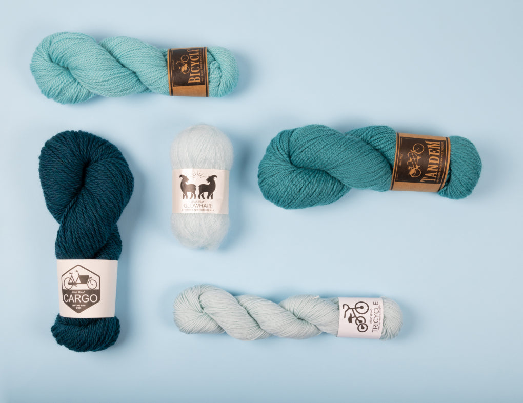 This image shows 5 skeins of yarn from the brand West Wool in blue gradients. Each skein is of a different West Wool base: Tandem, Tricycle, Glowhair, Bicycle, and Cargo. The skeins are placed on a light blue background. 