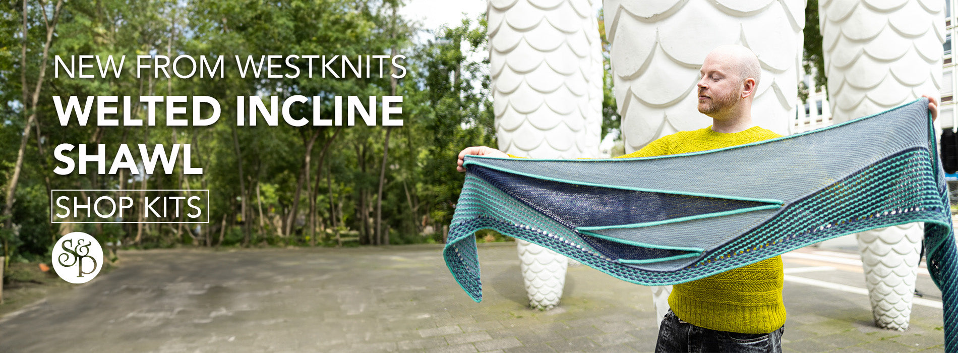 This image advertises the realise of Westknits new pattern called "Welted Incline Shawl". The text include a "shop kits" button and the black S&P logo. The image shows Stephen West wearing the Welted Incline Shawl. The shawl is knitted with blue tones. The background shows white pine-like structures and trees. 