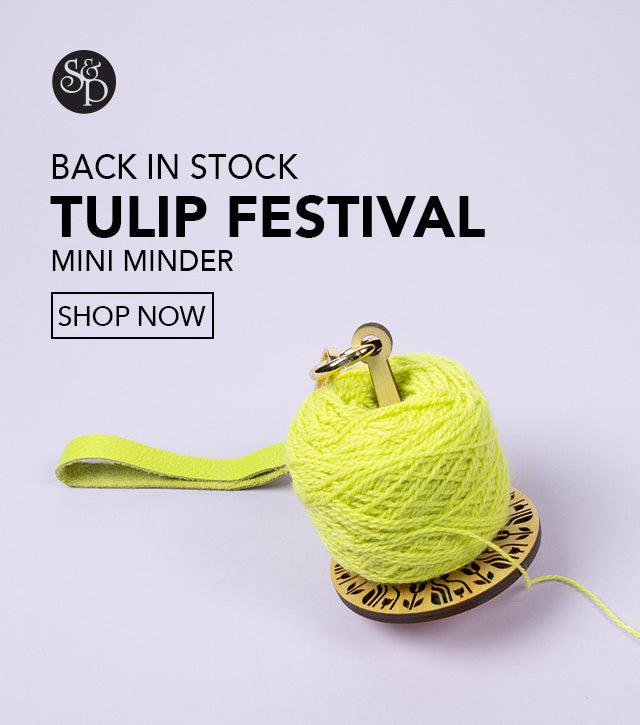 This image advertises that the "Tulip Festival" mini minder from the brand Lemonwood is back in stock at Stephen & Penelope. This mini minder is exclusive from Stephen & Penelope. The text includes a "shop now" button and the S&P black logo. The image shows a wooden mini minder with tulip decorations and a ball of neon yellow yarn on a light purple background. 