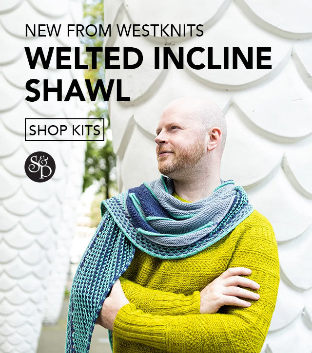 This image advertises the realise of Westknits new pattern called "Welted Incline Shawl". The text include a "shop kits" button and the black S&P logo. The image shows Stephen West wearing the Welted Incline Shawl. The shawl is knitted with blue tones. The background shows white pine-like structures. 